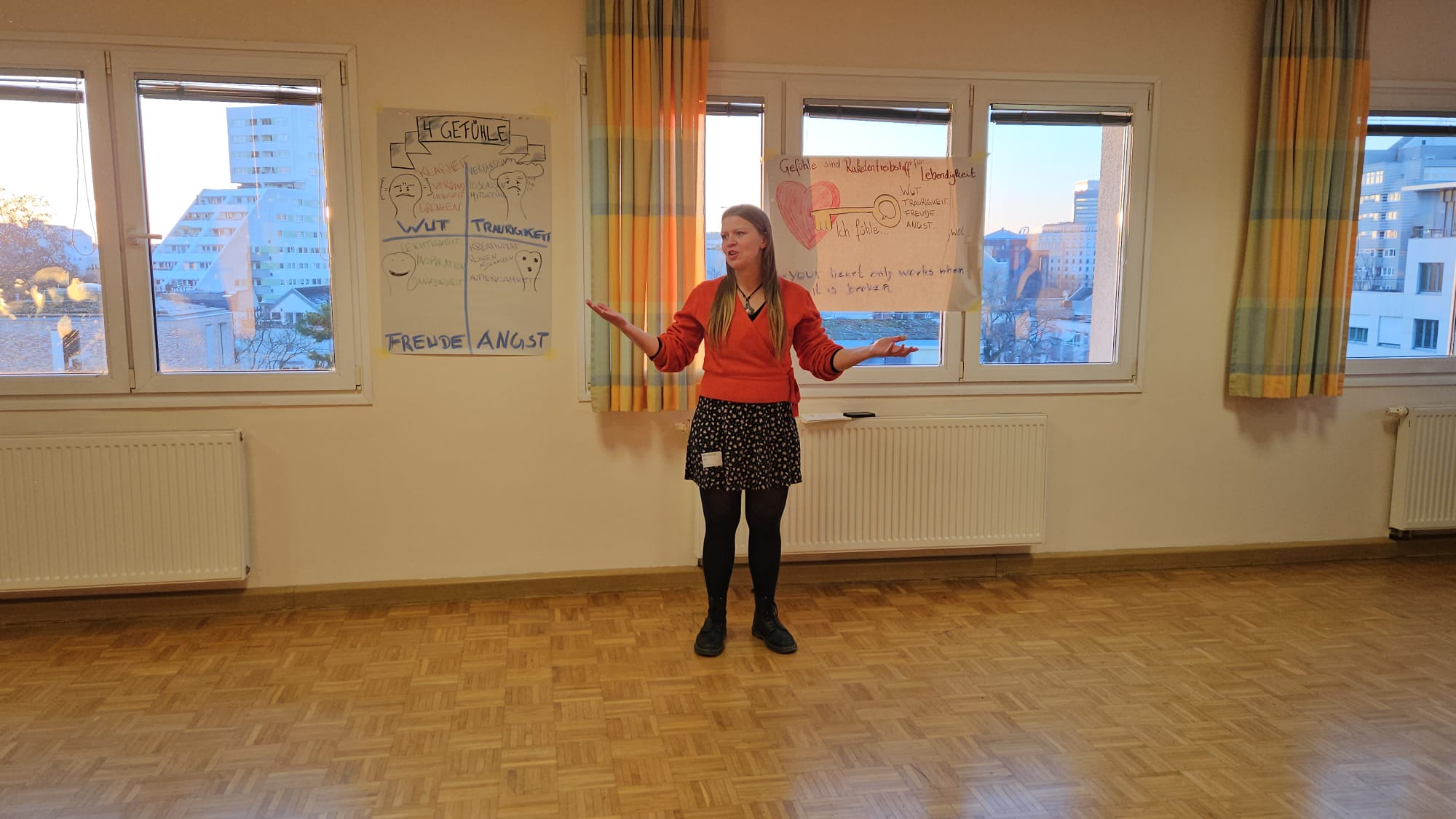 Thea Kupler steht in einem hellen Raum vor Notizen zu Gefühlen und spricht mit offenen Händen.